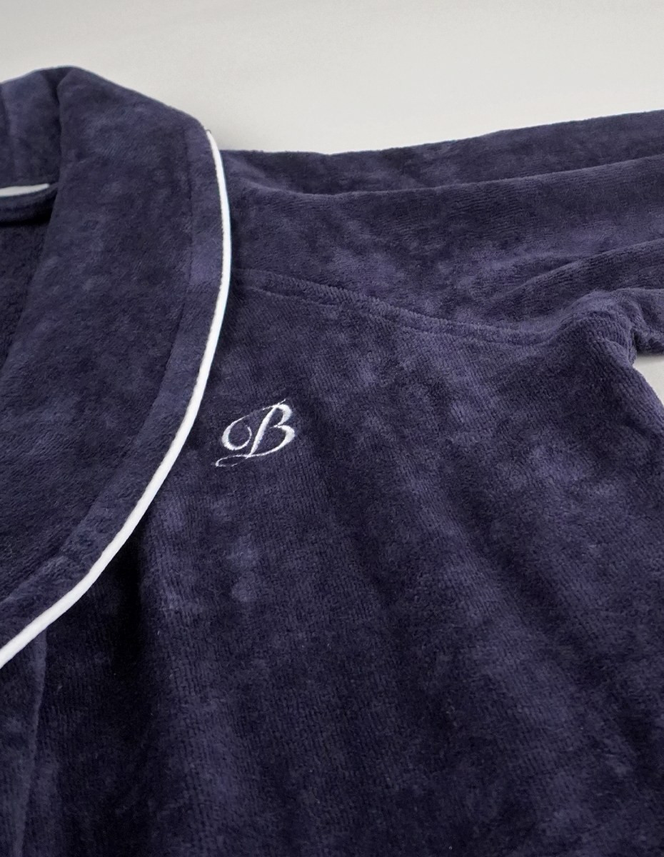 balmuir_portofino_robe_darknavy_closeup_2_web Der Bademantel Portofino besteht aus 100 % Baumwolle. Das Bademantel-Design zeichnet sich durch einen markanten Schalkragen und einen passenden Gürtel aus. Es ist mit Paspeldetails am Schalkragen und an den Ärmeln sowie einem raffiniert gestickten Markenlogo versehen. Unsere Portofino-Bademäntel werden in Portugal in perfekter Handarbeit gefertigt.